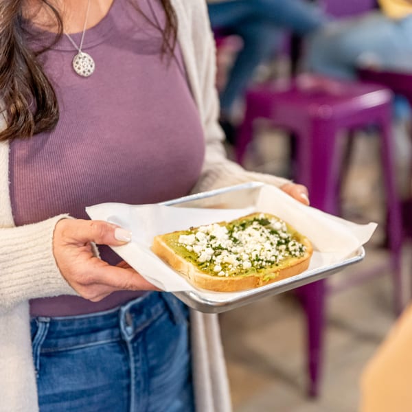 A person in a purple shirt and jeans holds a tray with avocado toast topped with feta.