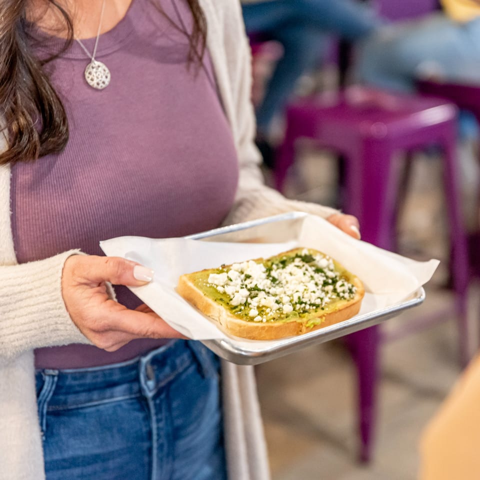 A person in a purple shirt and jeans holds a tray with avocado toast topped with feta.