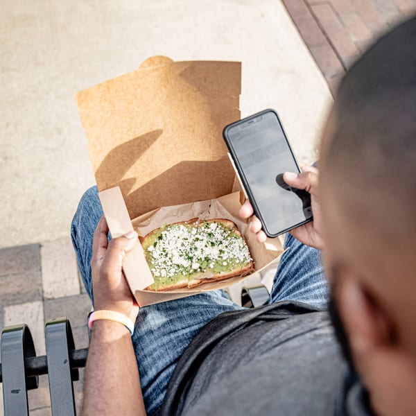 Person in jeans holding a Superfood Cafe avocado toast with feta and a smartphone outdoors.
