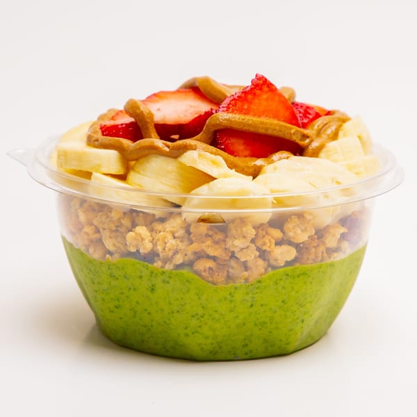 A Superfood Cafe green smoothie bowl with granola, bananas, strawberries, and peanut butter drizzle.