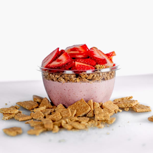 A Superfood Cafe strawberry smoothie bowl with granola and fresh strawberries, surrounded by cereal pieces.