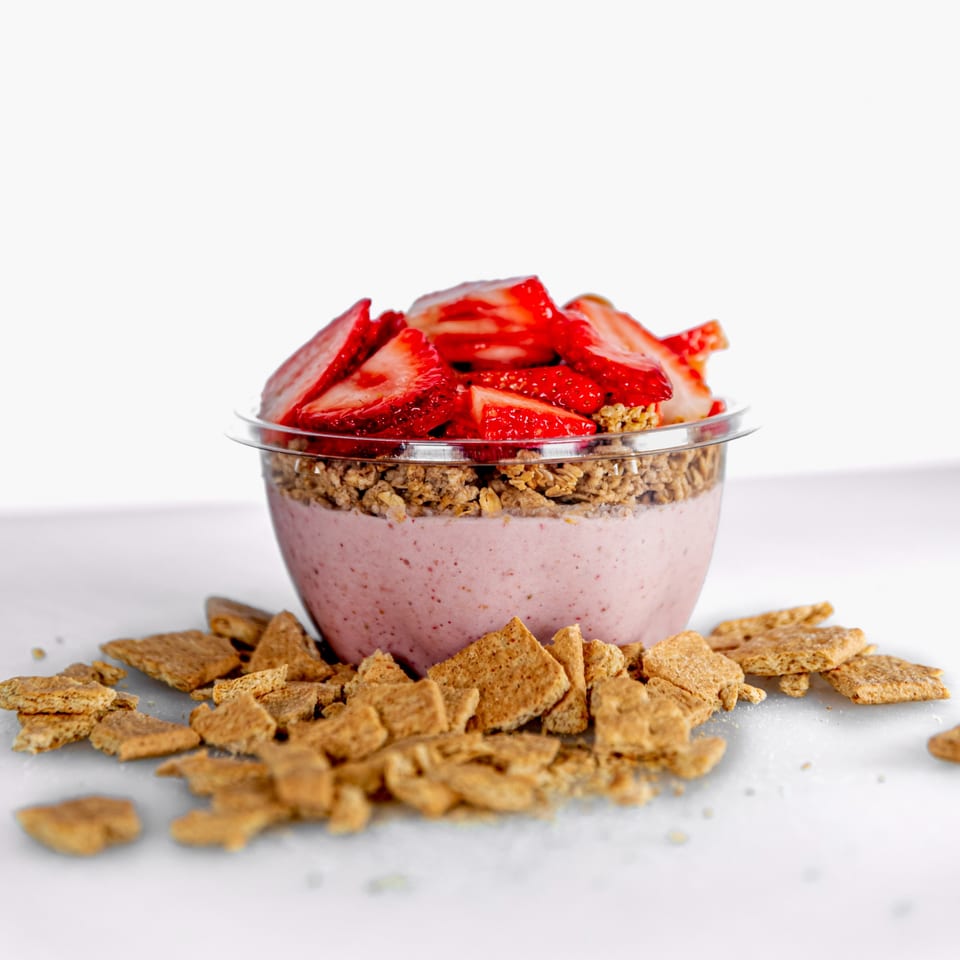 A Superfood Cafe strawberry smoothie bowl with granola and fresh strawberries, surrounded by cereal pieces.