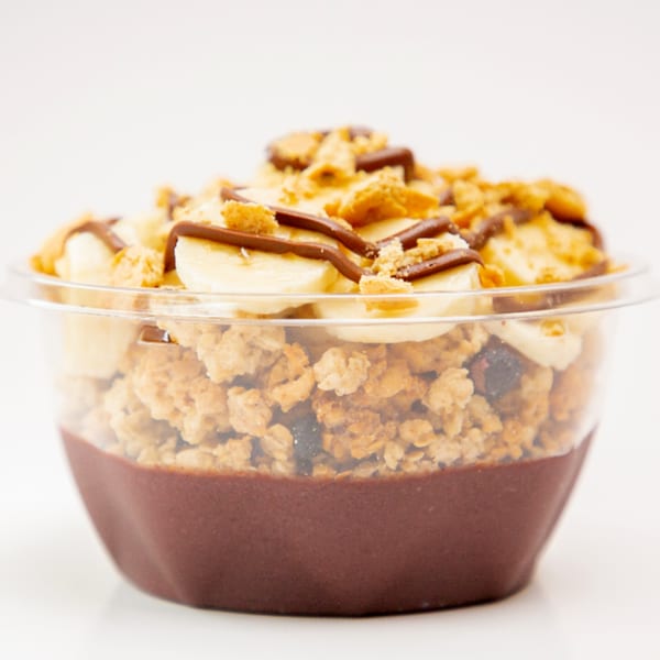 A Superfood Cafe acai bowl with granola, banana slices, and chocolate drizzle in a clear cup.