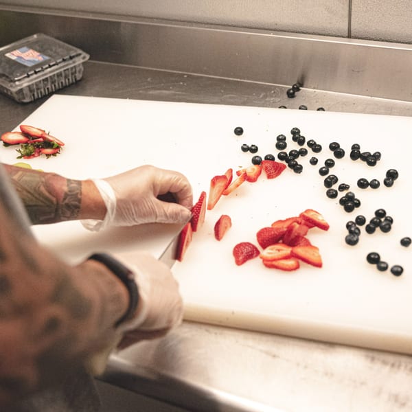 A chef in gloves slices fresh strawberries on a white cutting board next to blueberries.