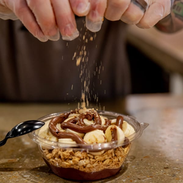 Gloved hands sprinkle granola over a Superfood Cafe acai bowl with bananas and chocolate drizzle.