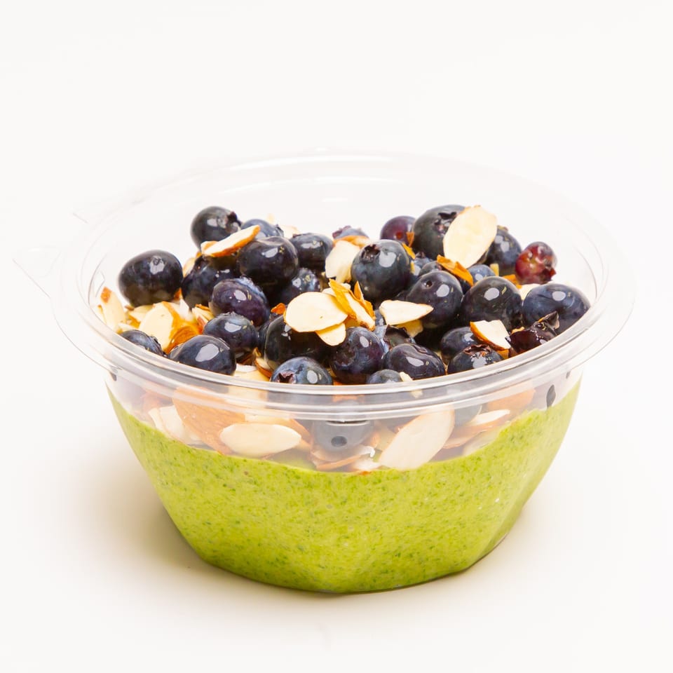 Green smoothie bowl topped with fresh blueberries and sliced almonds in a clear plastic container.