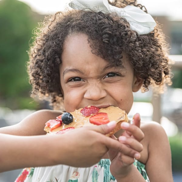 A young girl with curly hair and a white bow happily eats a Superfood Cafe fruit pizza.