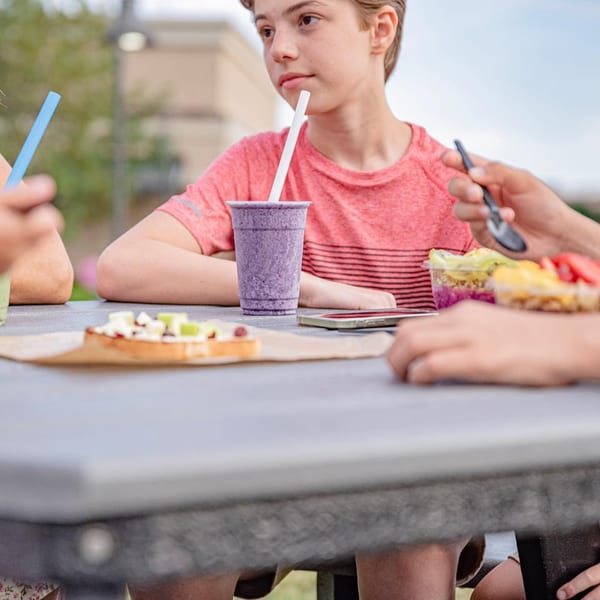 A boy sips a purple smoothie at an outdoor table with healthy Superfood Cafe items.