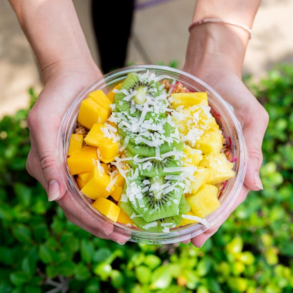 Hands holding a Superfood Cafe bowl with mango, kiwi, pineapple, and shredded coconut.