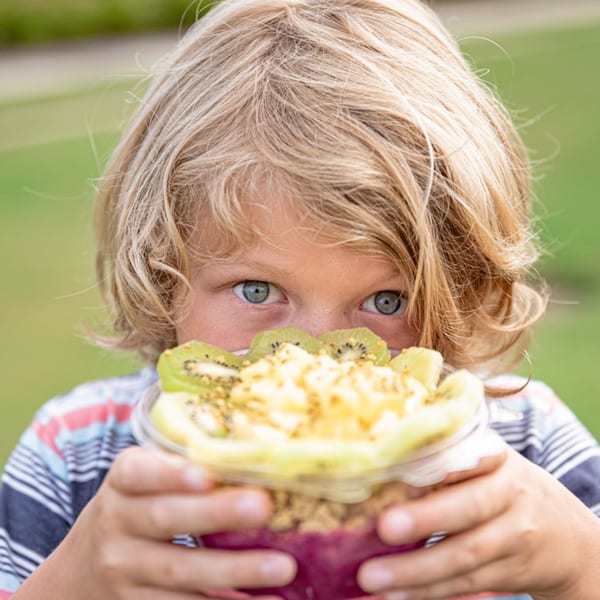 A child with blonde hair holds a Superfood Cafe acai bowl topped with kiwi and granola.