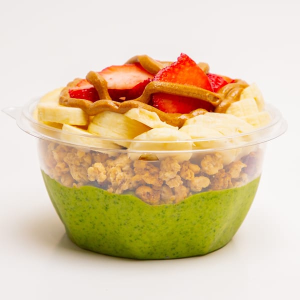 A Superfood Cafe green smoothie bowl with granola, banana, strawberries, and peanut butter drizzle.