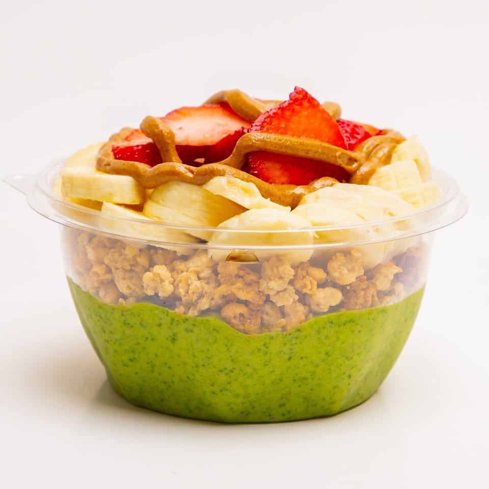 A Superfood Cafe green smoothie bowl with granola, banana, strawberries, and peanut butter drizzle.